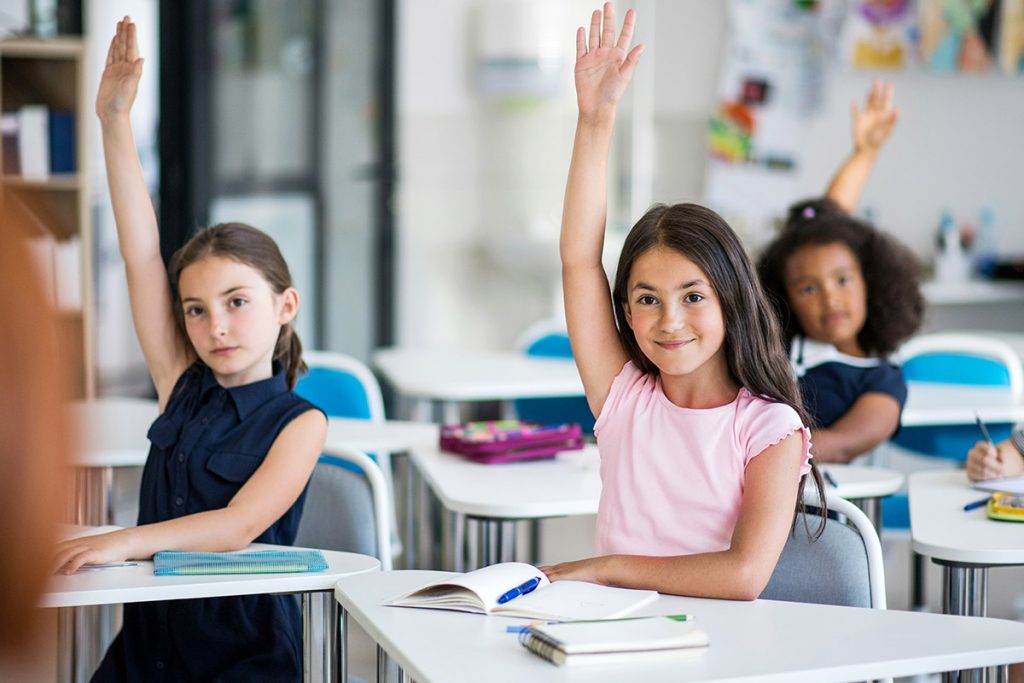 young-children-in-classroom-raising-their-hands-smiling